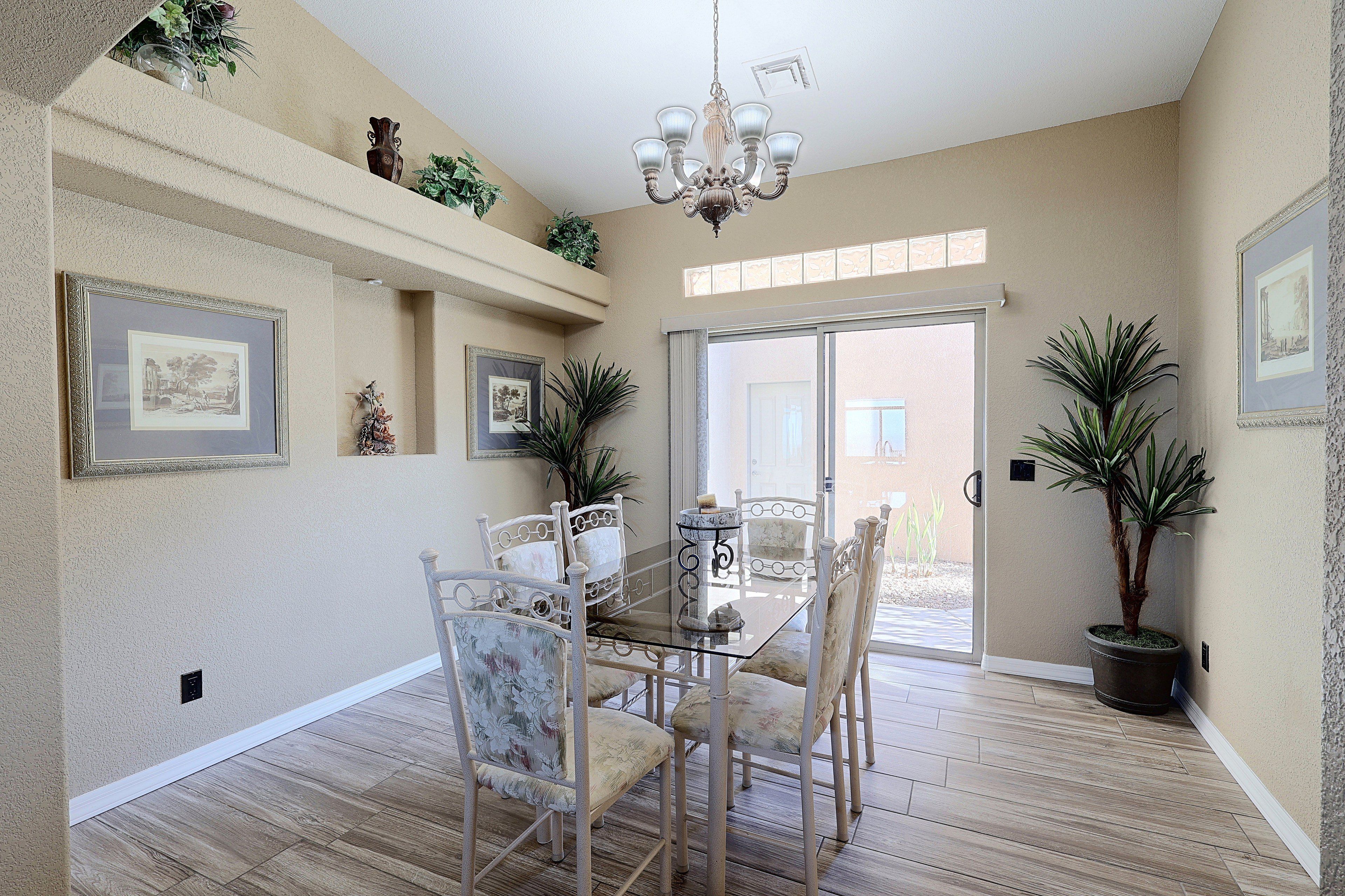 A dining area with a glass table, chairs, and a chandelier. There are framed pictures on the walls and potted plants.