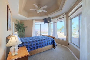 Une chambre d’hôtel équipée d’un lit, d’un bureau avec une lampe, d’un ventilateur de plafond et de grandes fenêtres offrant une vue sur des palmiers et un paysage lointain.