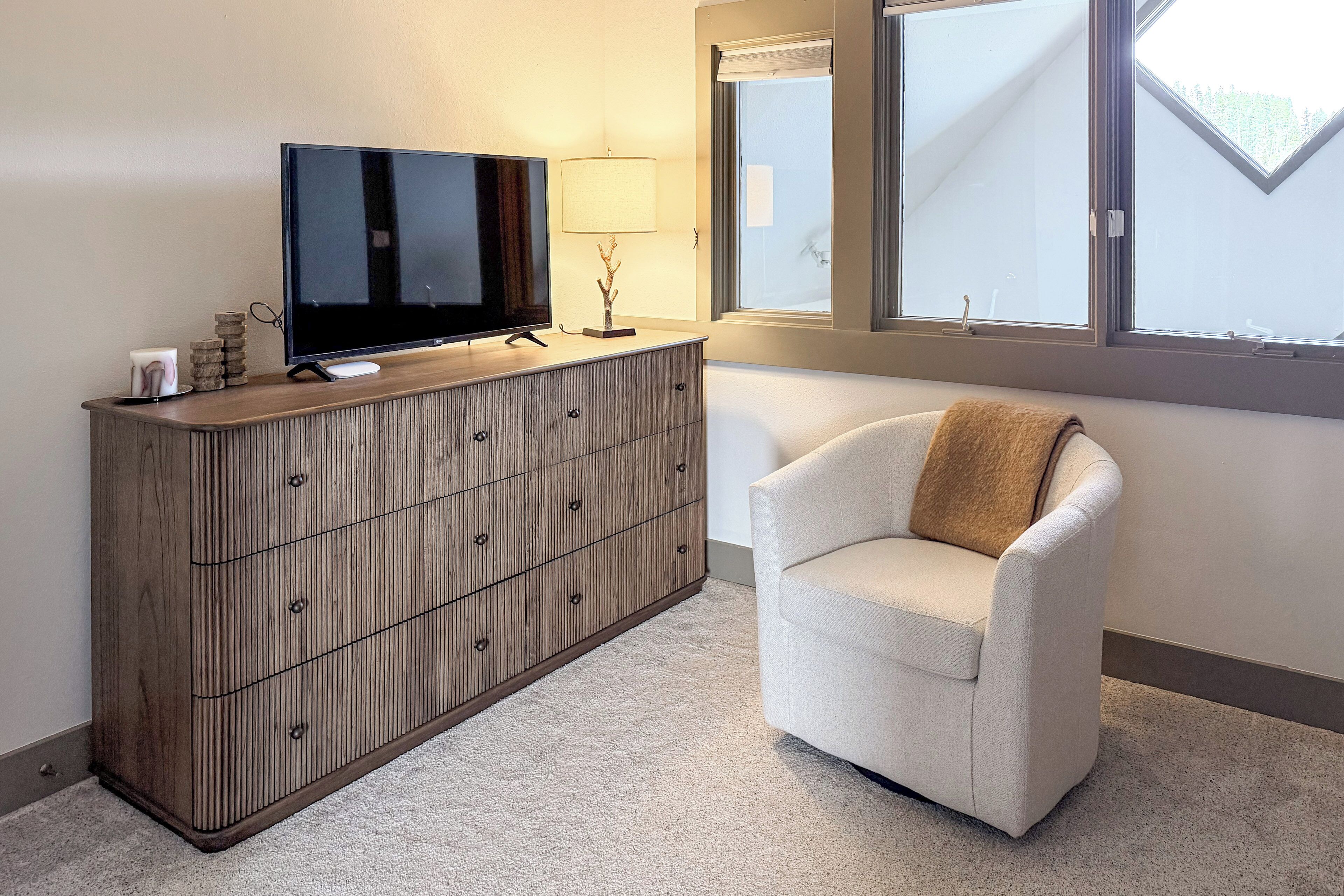 Une pièce avec un meuble en bois, une télévision, une lampe, une chaise et une fenêtre donnant sur un beau paysage.