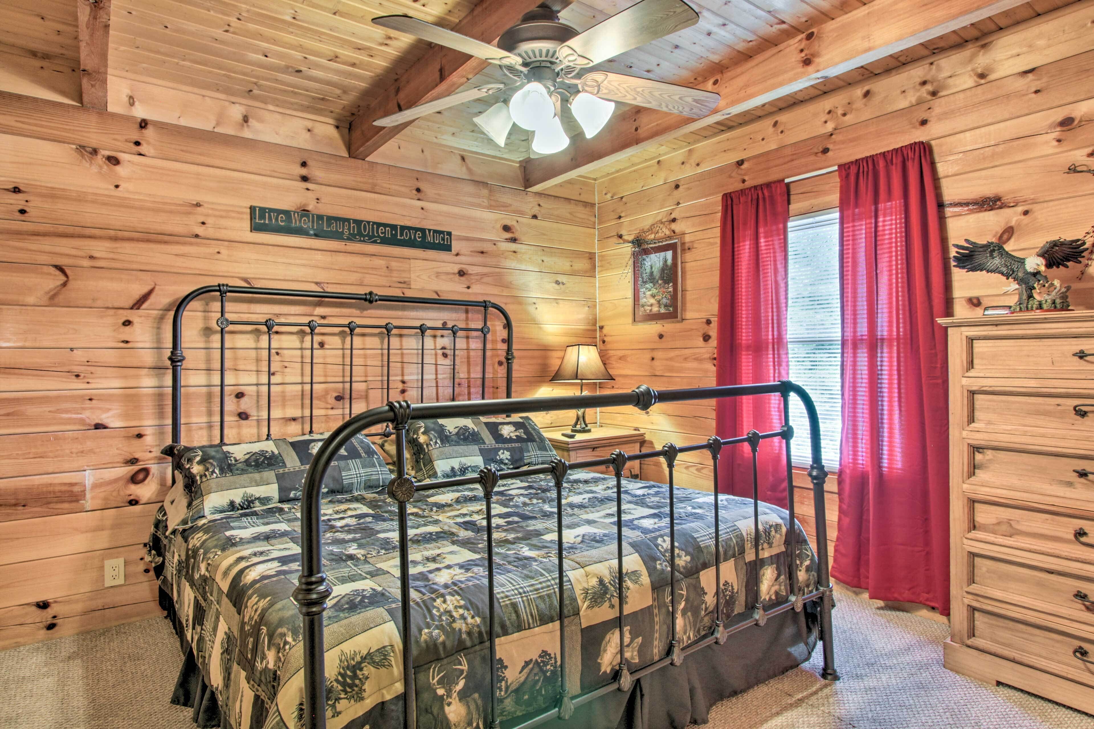 A wooden cabin bedroom with a metal bed frame, a wooden dresser, a ceiling fan, and a window with red curtains.