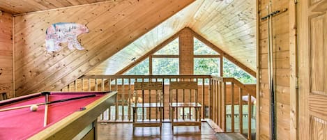 A wooden interior with a pool table, a bear mural, and a balcony with a view of trees.