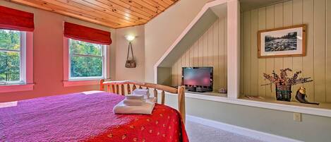 Un dormitorio con techo de madera, una cama con colcha roja, un televisor en una estantería y una ventana con vistas a árboles.