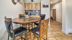 Un coin repas avec une table ronde en bois et quatre chaises, une cuisine avec des meubles en bois et un dosseret carrelé, et un couloir menant à une porte en bois.