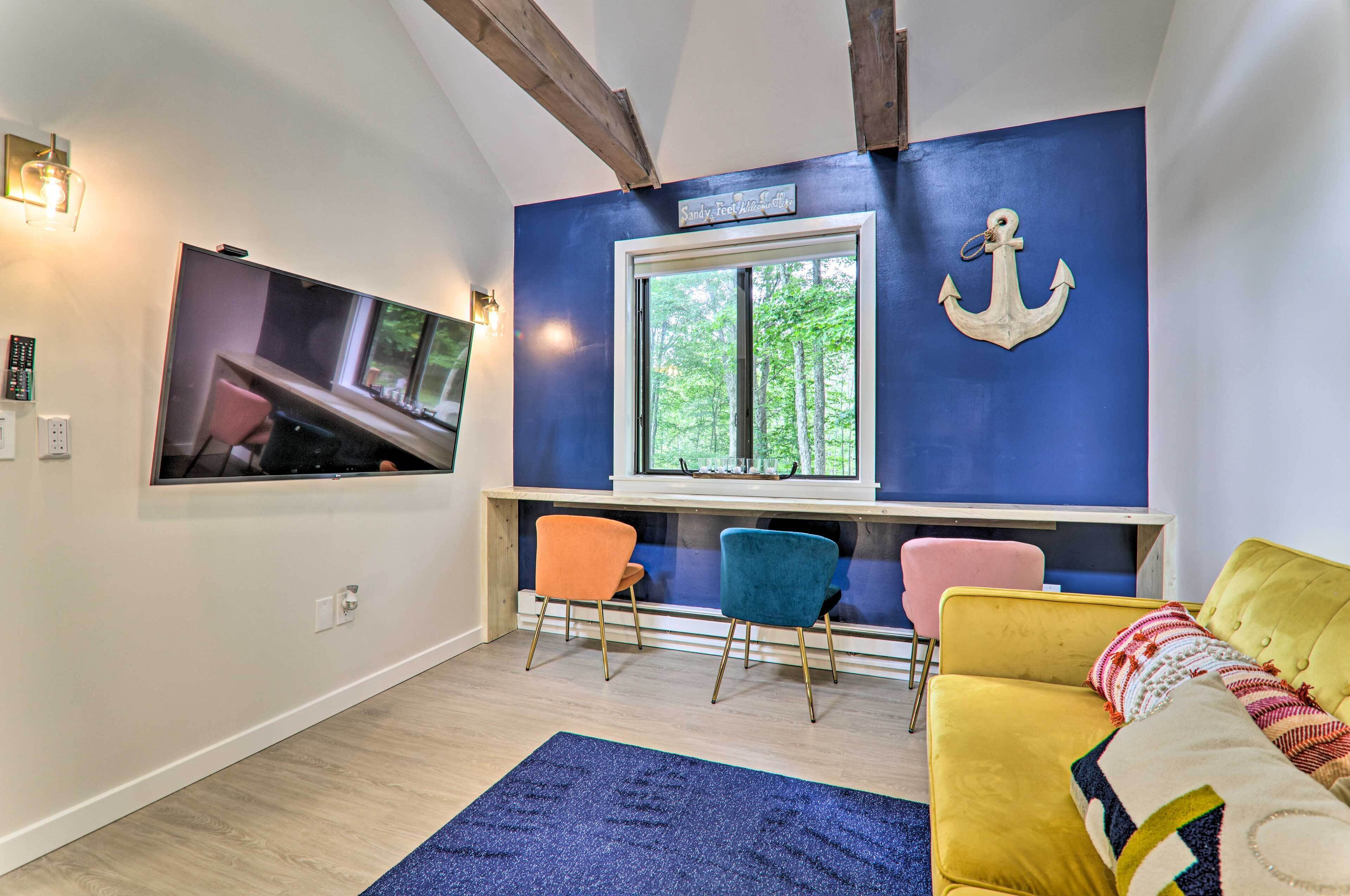 A room with a large TV, a window with a view of trees, a yellow sofa, and colorful chairs.