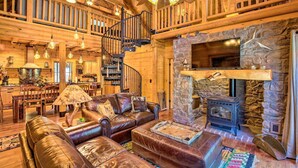 A spacious log cabin interior with a stone fireplace, leather furniture, a wooden staircase, and a large TV mounted on the wall. - Luxury Mountain Cabin w/ Furnished Deck + Views! (Mount Lemmon)