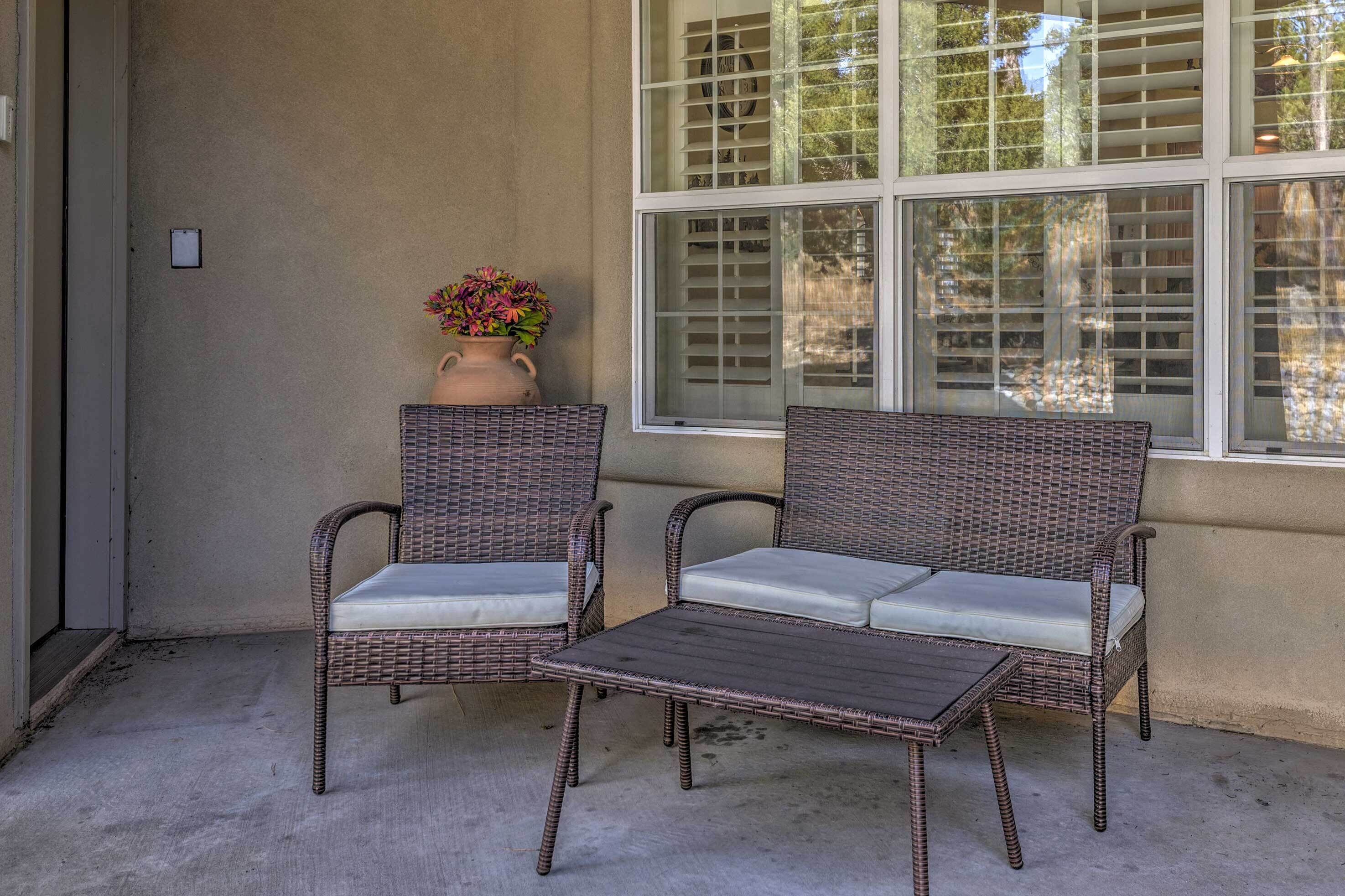 Un sofá y sillas de mimbre con cojines, una mesita y un jarrón con flores en un patio de concreto.