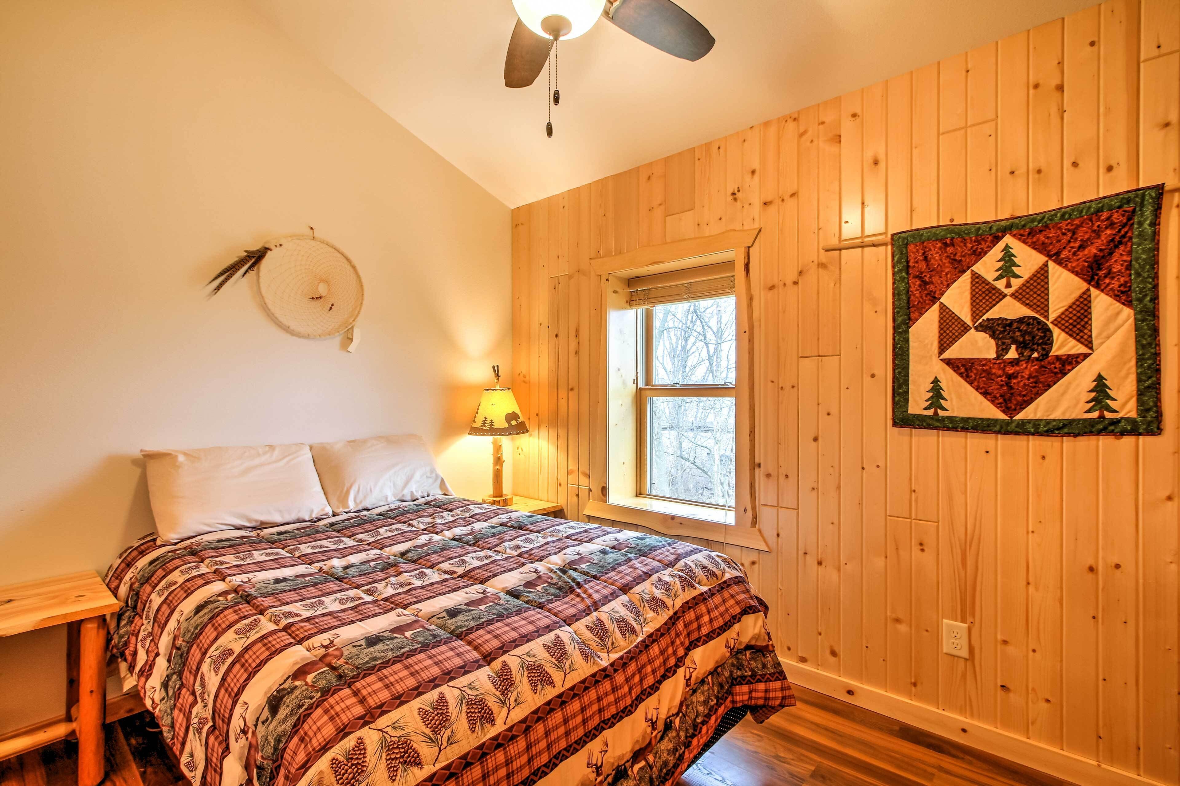 A bedroom with a wooden floor, a bed with a quilt, a window, a wall clock, and a quilted wall hanging.