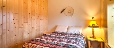 A wooden-paneled room with a bed, a wall-mounted dreamcatcher, and a small table with a lamp.