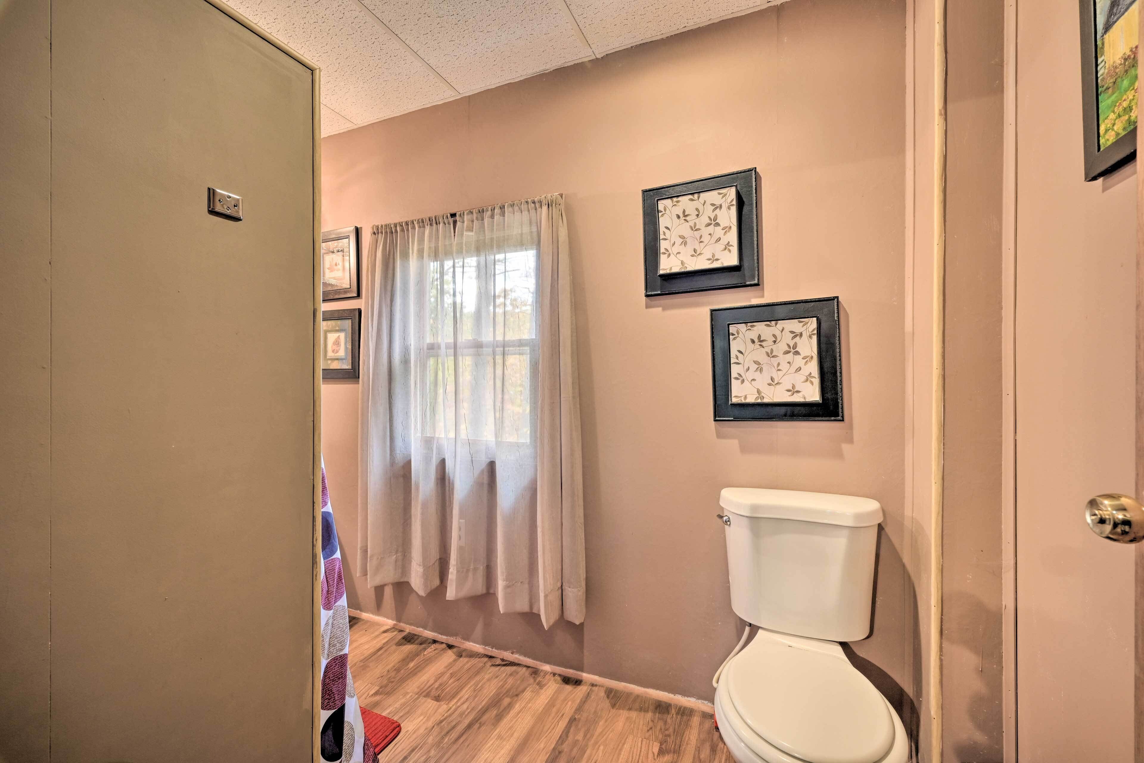 A bathroom with a toilet, wooden flooring, a window with a sheer curtain, and two framed pictures on the wall.