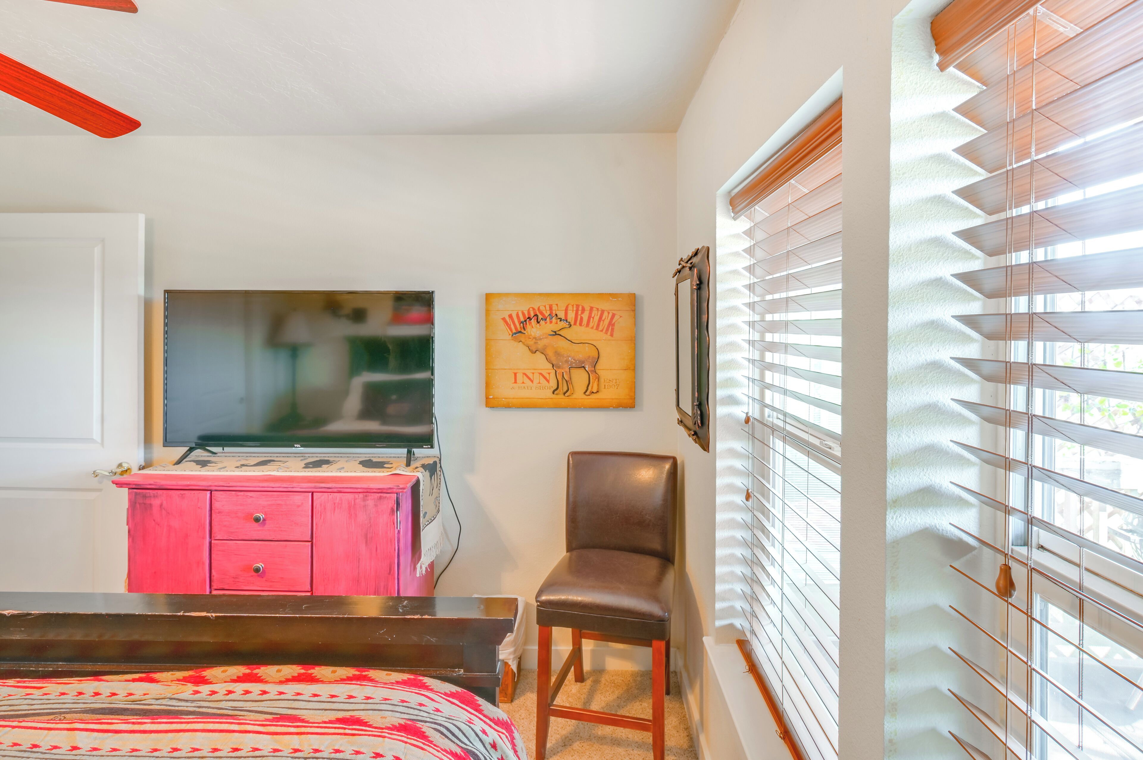 A bedroom with a bed, a pink dresser, a chair, a television, and a wall-mounted picture of a moose.