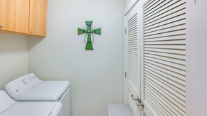 A laundry room with two white washing machines, a wooden cabinet, a green cross decoration, and a white door with horizontal blinds. - Walk to Main St! Ennis Townhome w/ Balcony (Ennis)