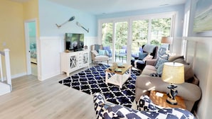 A living room with a large TV, a coffee table, and a sofa. There are windows with a view of greenery outside. - 2 Mi to Bethany Beach: Family-friendly Home! (Ocean View)