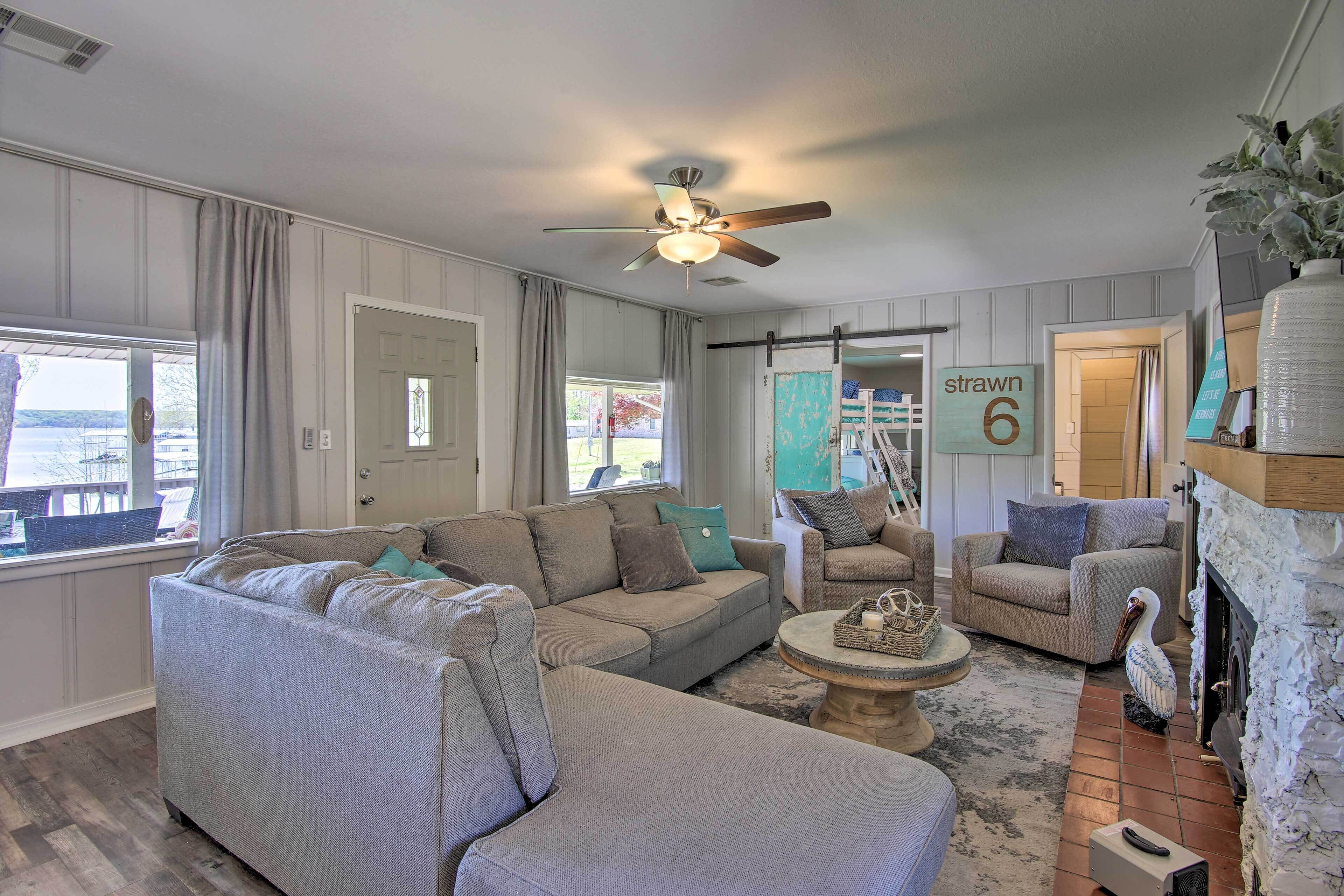 A living room with a sectional sofa, a round coffee table, and a fireplace. There is a ceiling fan and a view of the outside through a window.