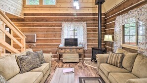 Ein gemütliches Blockhaus-Interieur mit einer Holztreppe, einem Fernseher auf einem Holztisch, einem Kamin und zwei Sofas mit gemusterten Kissen.
