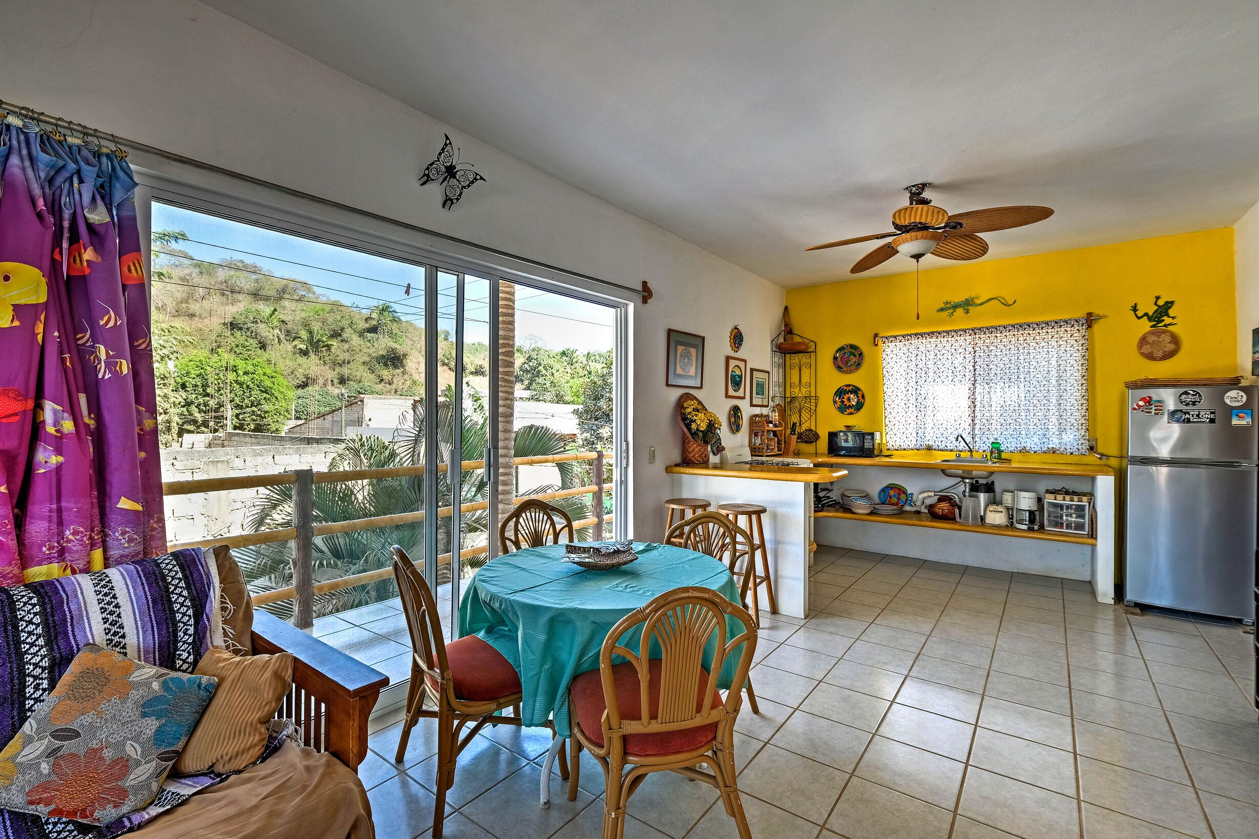 A bright kitchen with a yellow wall, a dining area with a table and chairs, and a balcony with a view of the hills.