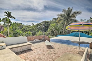 A rooftop pool area with a colorful umbrella, lounge chairs, and a view of lush greenery.