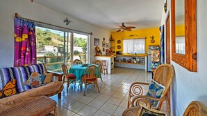 A spacious living area with a sofa, dining table, and chairs, a kitchen area with a counter and appliances, and large windows offering a view of the outdoors. - Sayulita Condo: Walk to Beach, Plaza & Dining! (Sayulita)