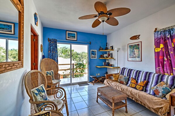 A room with a ceiling fan, wicker furniture, a sofa with colorful cushions, and a view through a glass door to the outside. - Sayulita Condo: Walk to Beach, Plaza & Dining! (Sayulita)