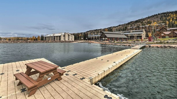 A wooden dock with a picnic table extending into a calm lake, surrounded by buildings with a mountainous backdrop. - Brian Head Condo w/ Patio & Community Amenities! (Brian Head)