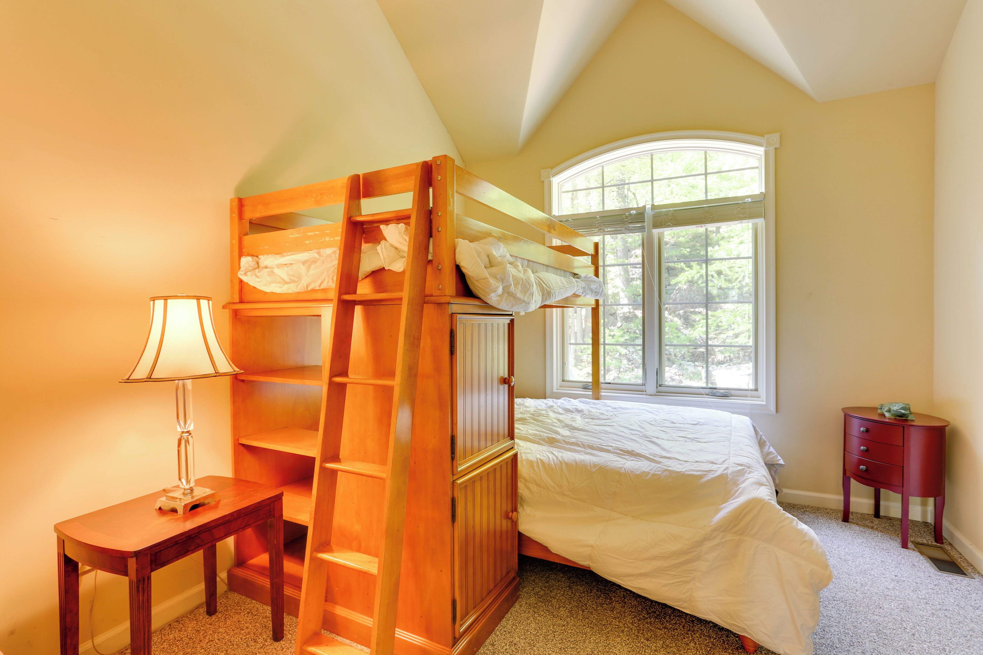 A bedroom with a bunk bed, a bedside table with a lamp, a window with a view of trees, and a small wooden nightstand.