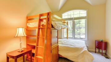 A bedroom with a bunk bed, a bedside table with a lamp, a window with a view of trees, and a small wooden nightstand.