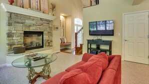 A living room with a stone fireplace, a glass coffee table, a red sofa, a television on a black stand, and a staircase leading to an upper level.
