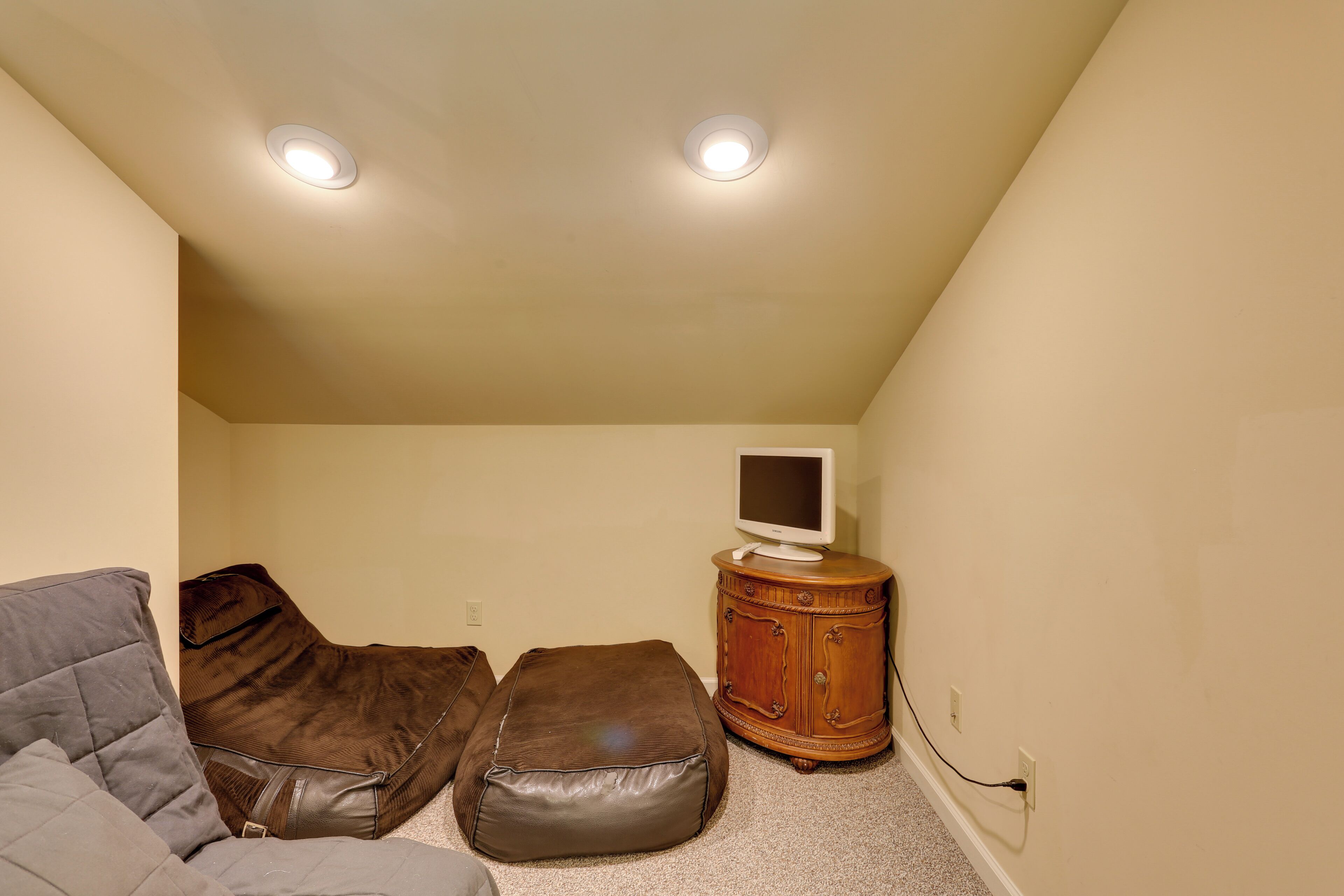 Una habitación pequeña y acogedora con un sofá gris, un reposapiés de cuero marrón, un mueble de madera con un televisor de pantalla plana y dos lámparas de techo.