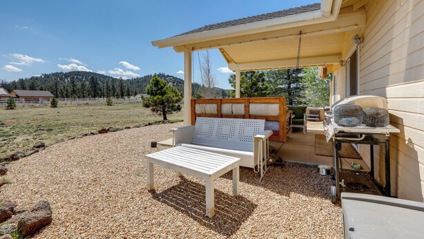 A covered patio area with a white table and bench, a grill, and a wooden fence in the background. - Flagstaff Home w/ Hot Tub & Mountain Views (Flagstaff)