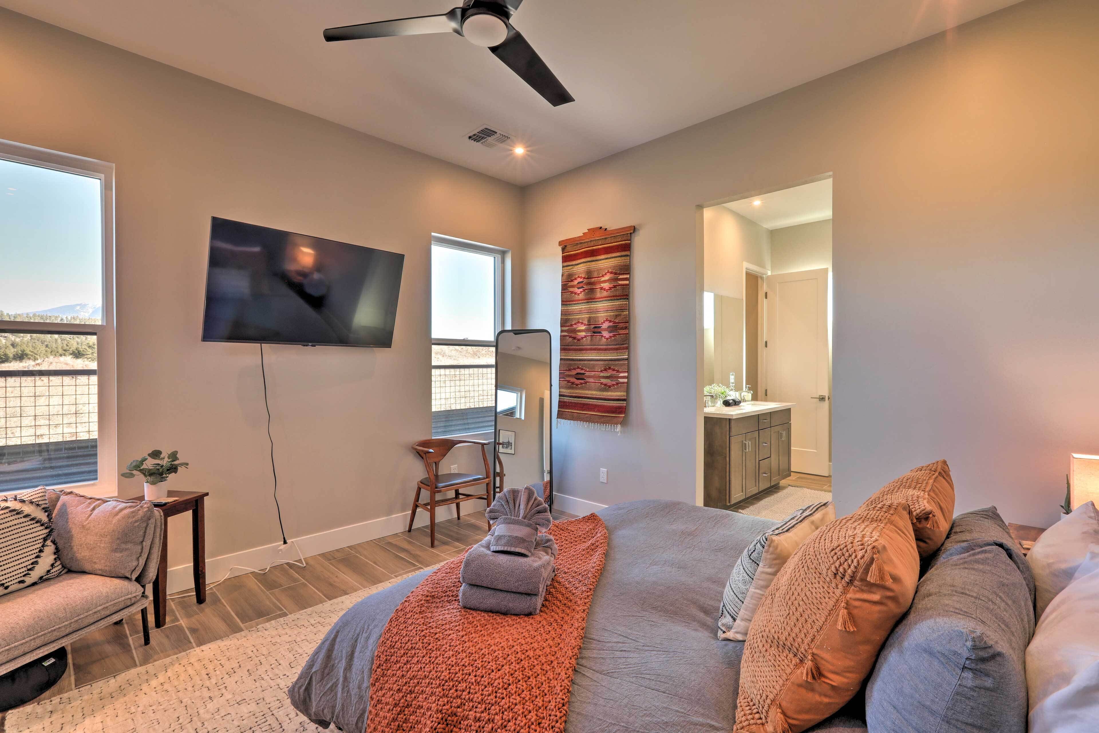 A modern bedroom with a large bed, a flat-screen TV mounted on the wall, a ceiling fan, and a view of the outdoors through a window.