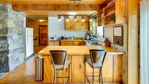 A wooden kitchen with a stone wall, wooden cabinets, and a bar area with two chairs. - New York Vacation Rental w/ Large Deck! (Saugerties)
