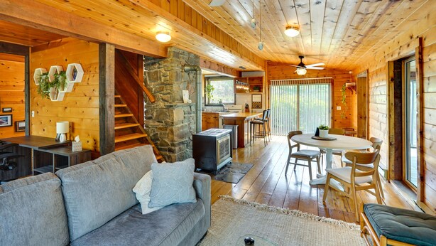 A cozy cabin interior with a stone fireplace, wooden walls, a dining area, and a kitchen. - New York Vacation Rental w/ Large Deck! (Saugerties)