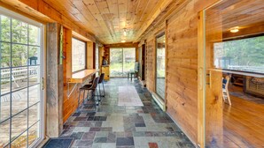 A wooden cabin interior with a stone-tiled floor, large windows, and a dining area with chairs and a table. - New York Vacation Rental w/ Large Deck! (Saugerties)