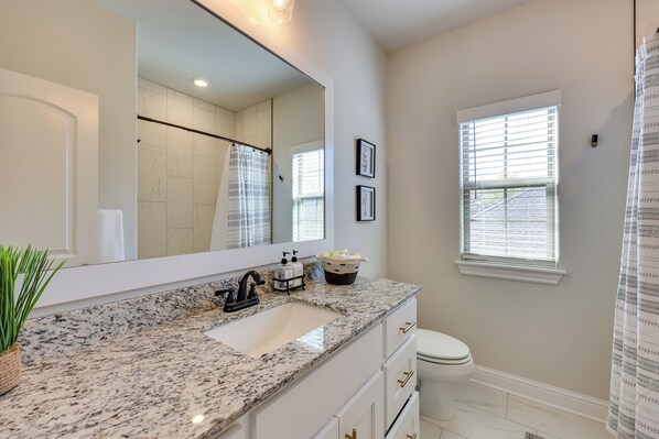 A bathroom with a granite countertop sink, a large mirror, a toilet, and a window with blinds.