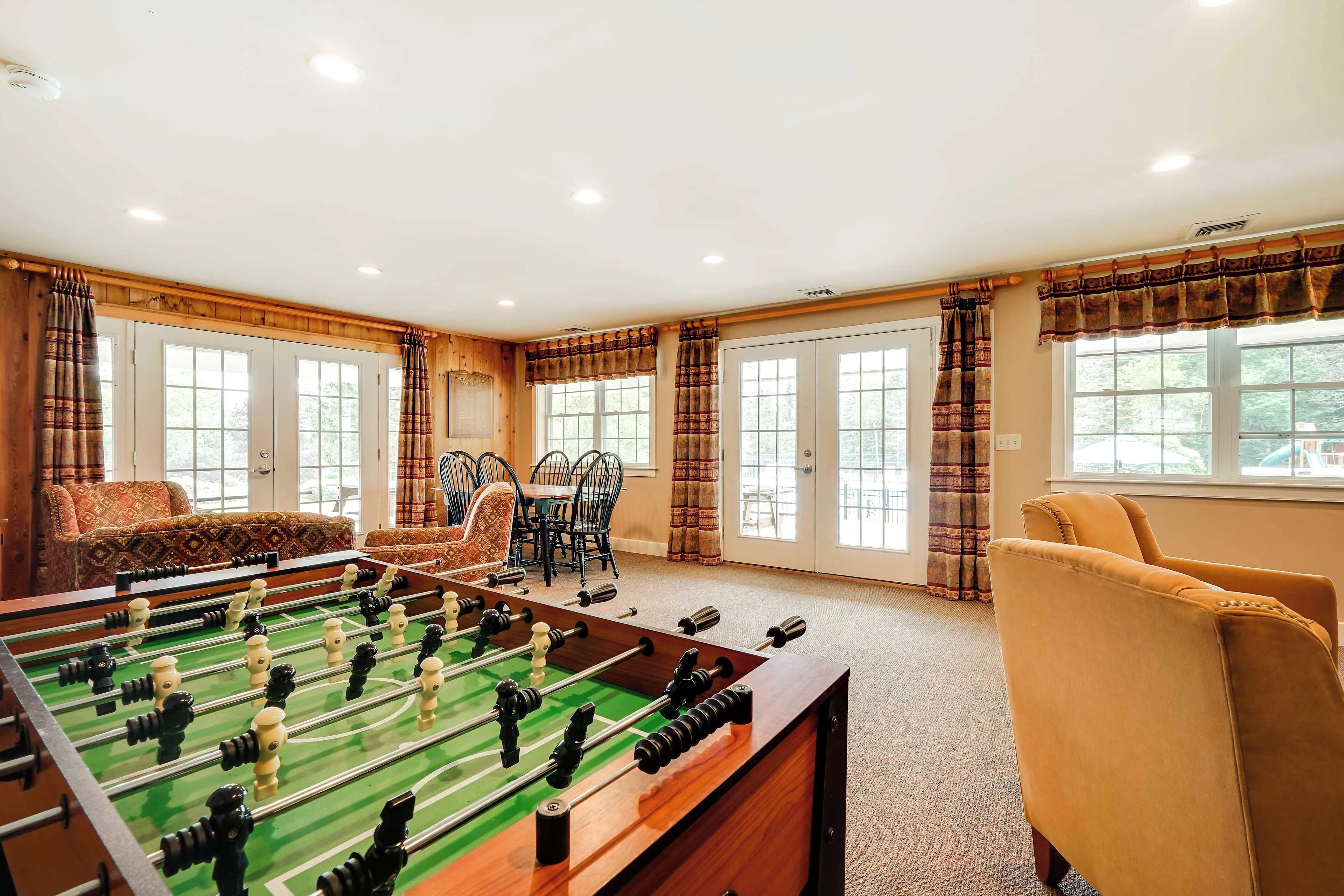 A room with a foosball table, a dining area with chairs and a table, and large windows with curtains.