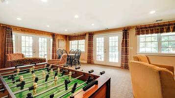 A room with a foosball table, a dining area with chairs and a table, and large windows with curtains.