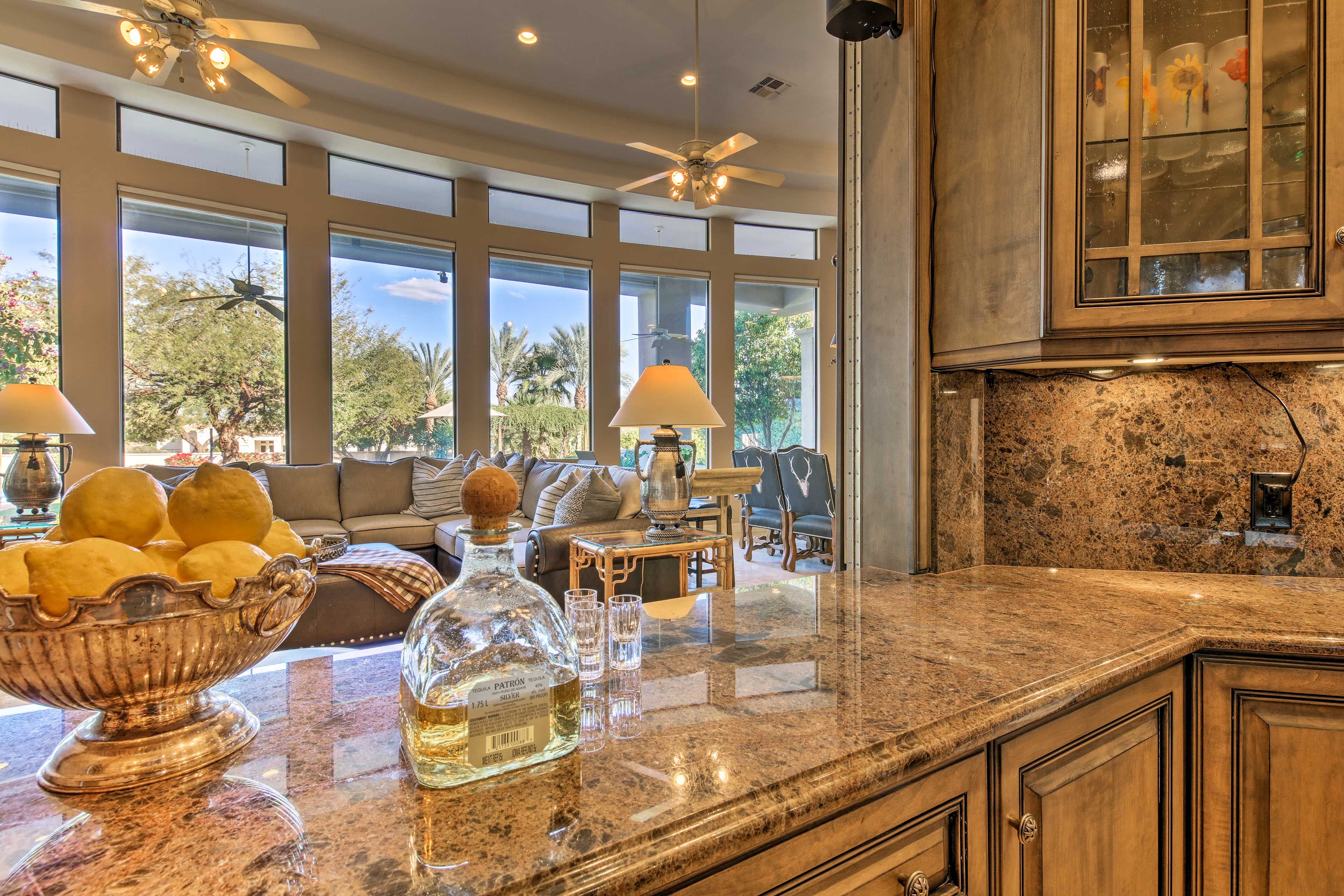 A kitchen with granite countertops, a large bowl of lemons, a bottle of liquor, and a dining area with a sofa and lamps.