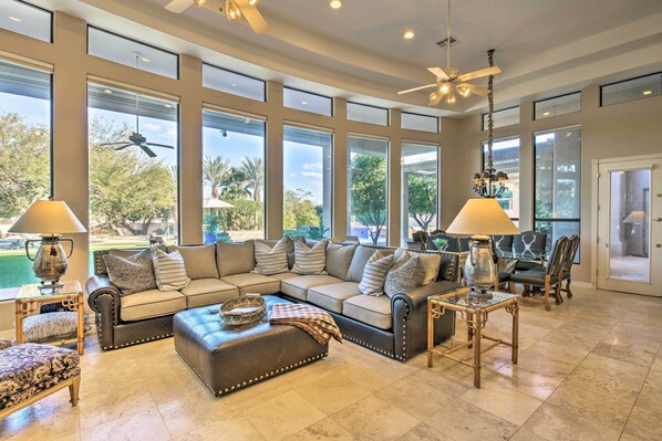 A spacious living room with a sectional sofa, ottoman, and two side tables with lamps. Large windows provide a view of the outdoors.