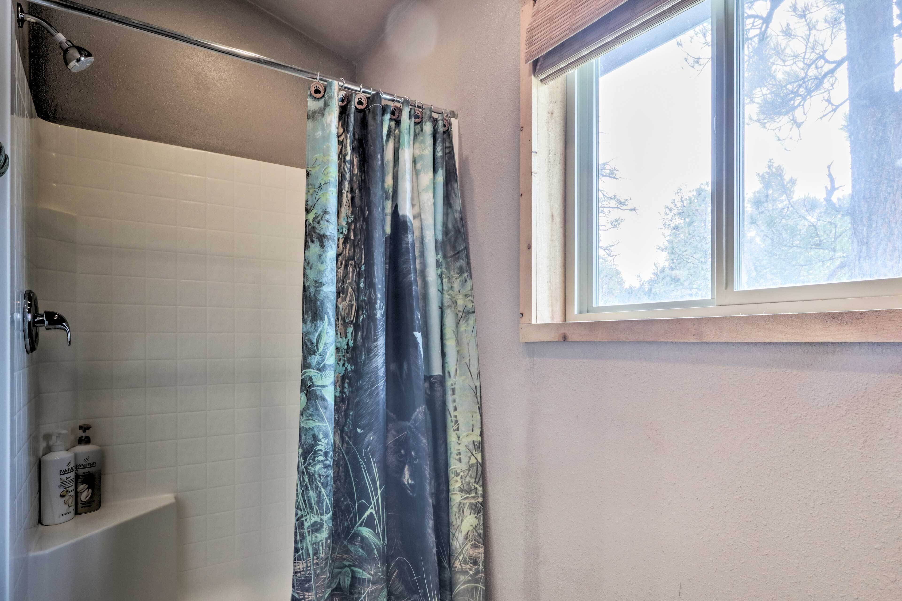 A bathroom with a shower curtain featuring a forest scene, a window with a view of trees, and a white tiled wall.