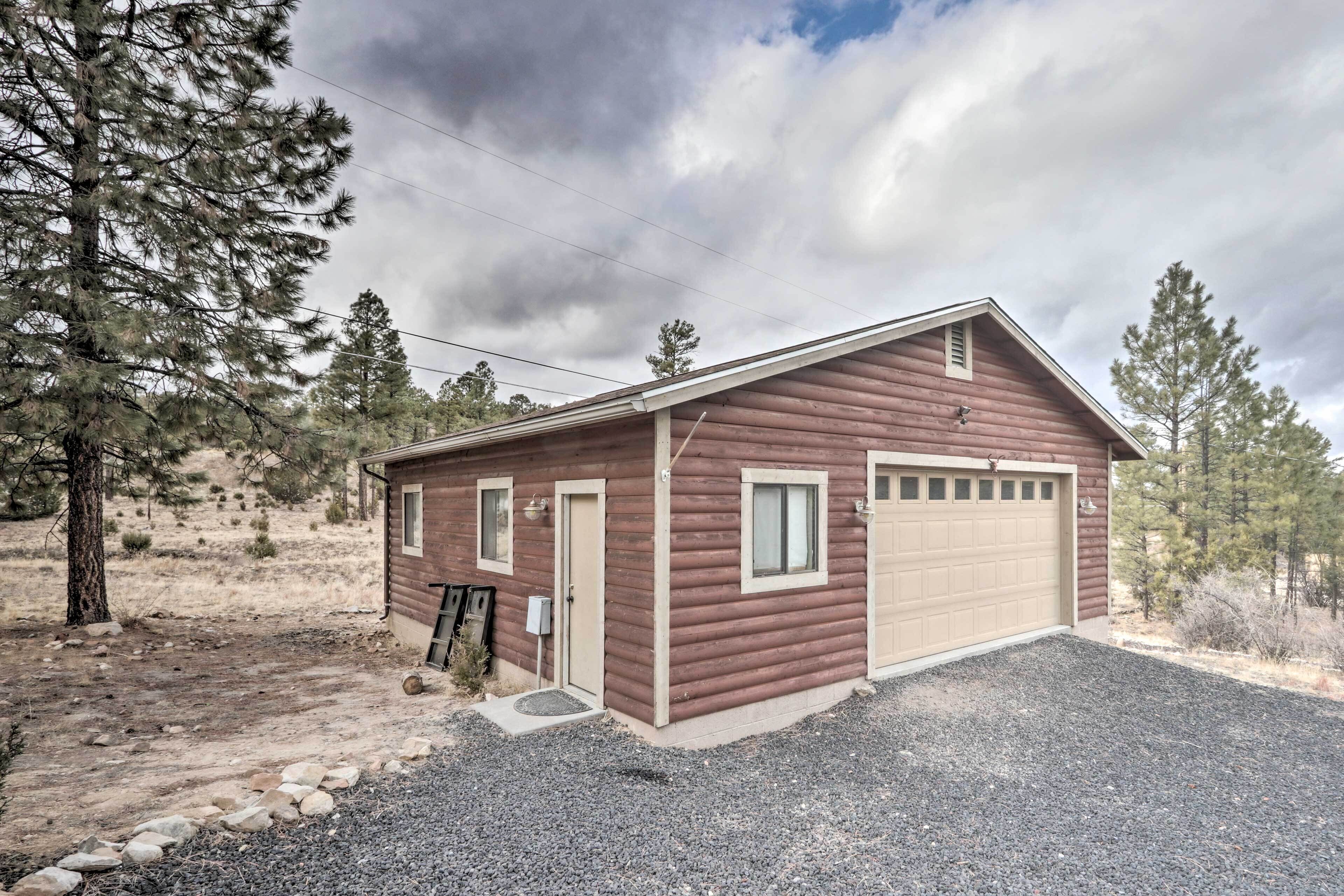 A single-story log cabin with a beige garage door, surrounded by a gravel driveway and sparse vegetation.