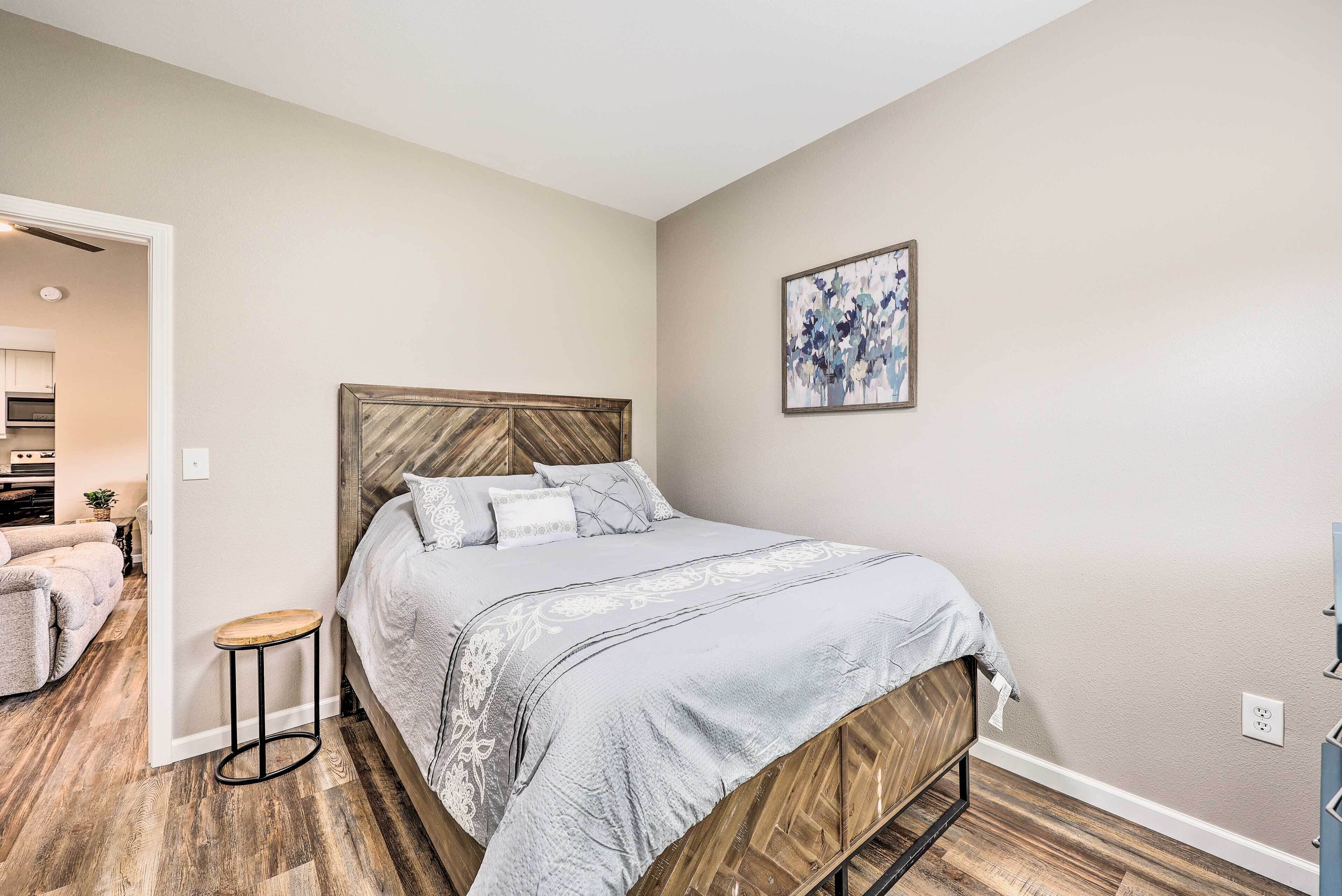 A bedroom with a bed, a small stool, a wooden headboard, a painting on the wall, and a kitchen visible through an open door.