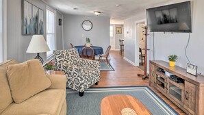 A living room with a sofa, a patterned armchair, a dining area with a table and chairs, a TV mounted on the wall, and a wooden cabinet with a 'Wi-Fi' sign. - Downtown Canandaigua Apt: 1 Mi to Lake & Pier! (Canandaigua)