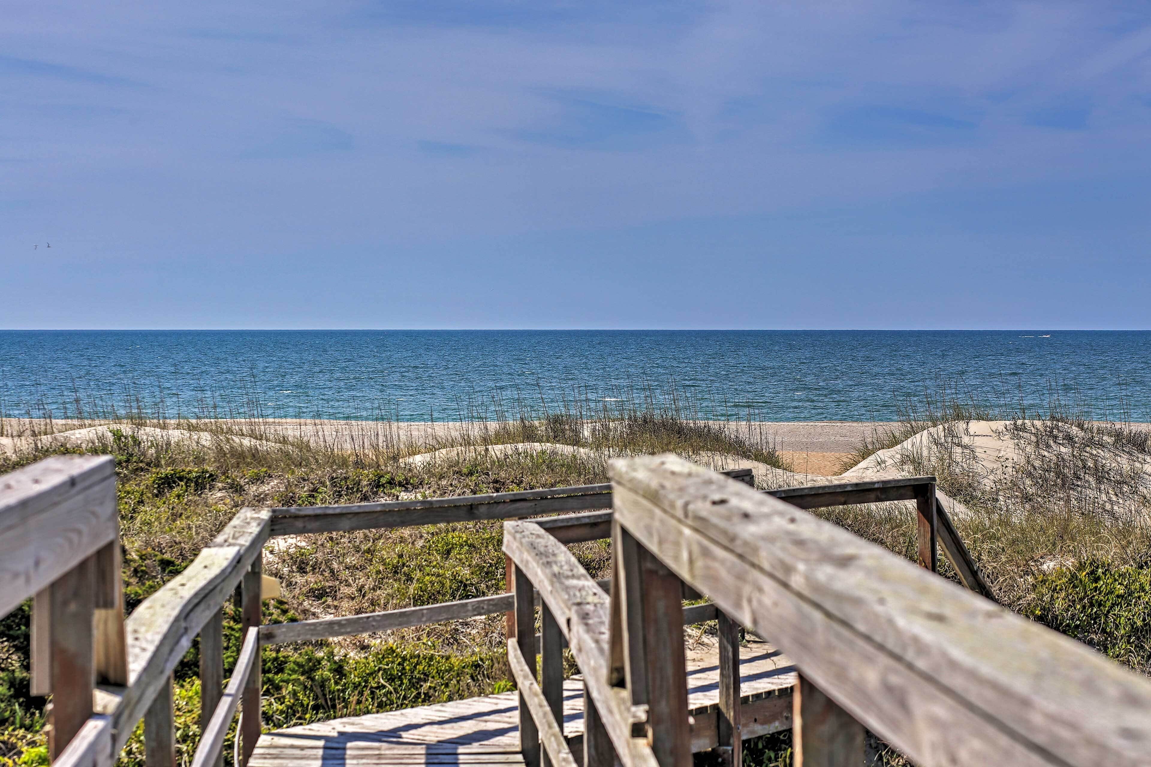 A wooden boardwalk leading to a beach with sand dunes and the ocean in the background.