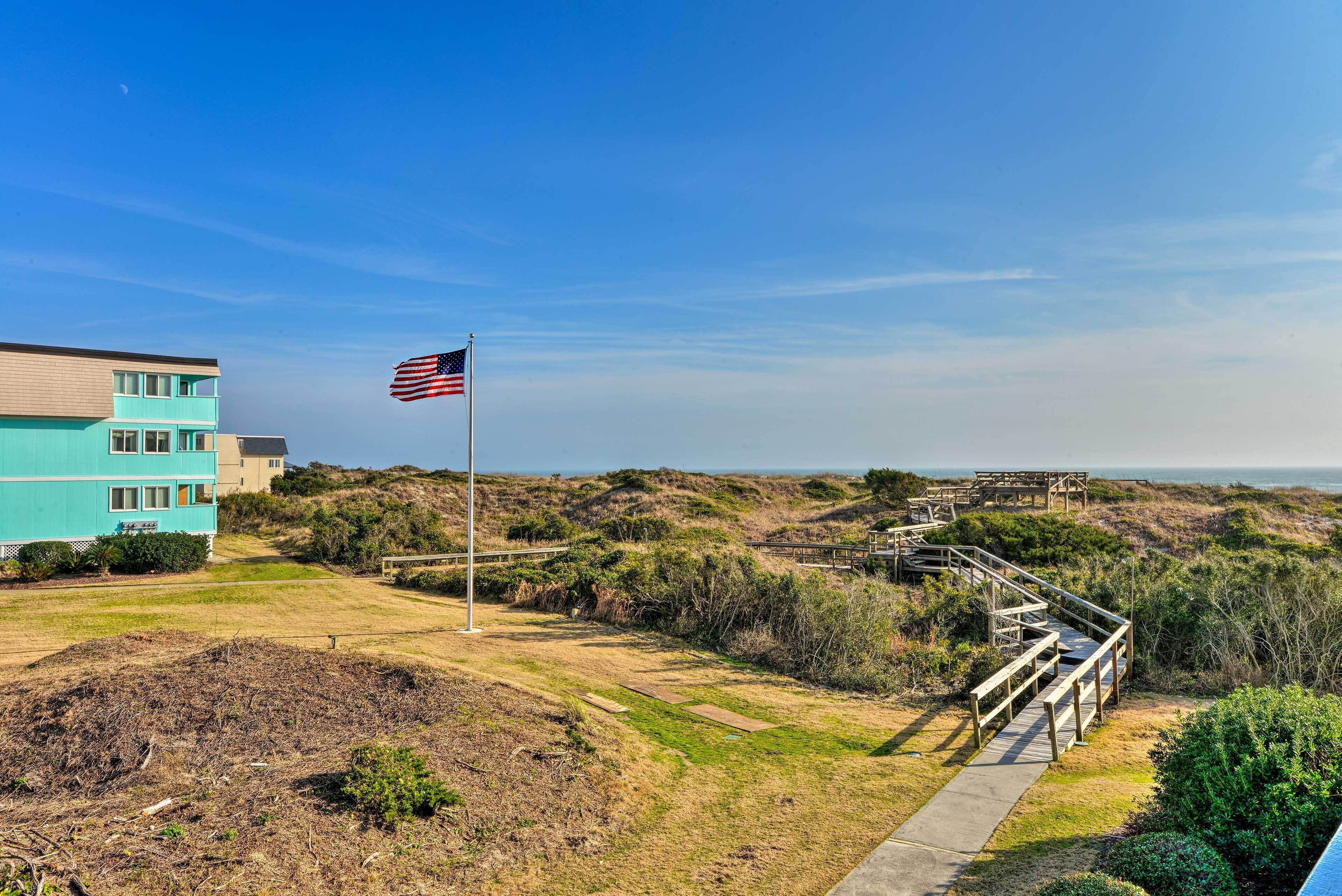 Un bâtiment situé sur la côte, doté d’un mât pour drapeau, d’une promenade en bois et offrant une vue sur la plage et les dunes.