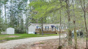 A single-story mobile home with a deck, surrounded by trees and a gravel driveway.