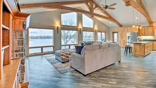 A spacious living room with a large couch, a stone accent wall, and a view of a snowy landscape through large windows. - Dock, Hot Tub & Game Room: Luxe Lake Latoka Home (Alexandria)