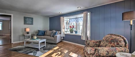 A living room with a sofa, armchair, and coffee table. There is a window with curtains, a lamp, and a wall-mounted sign.