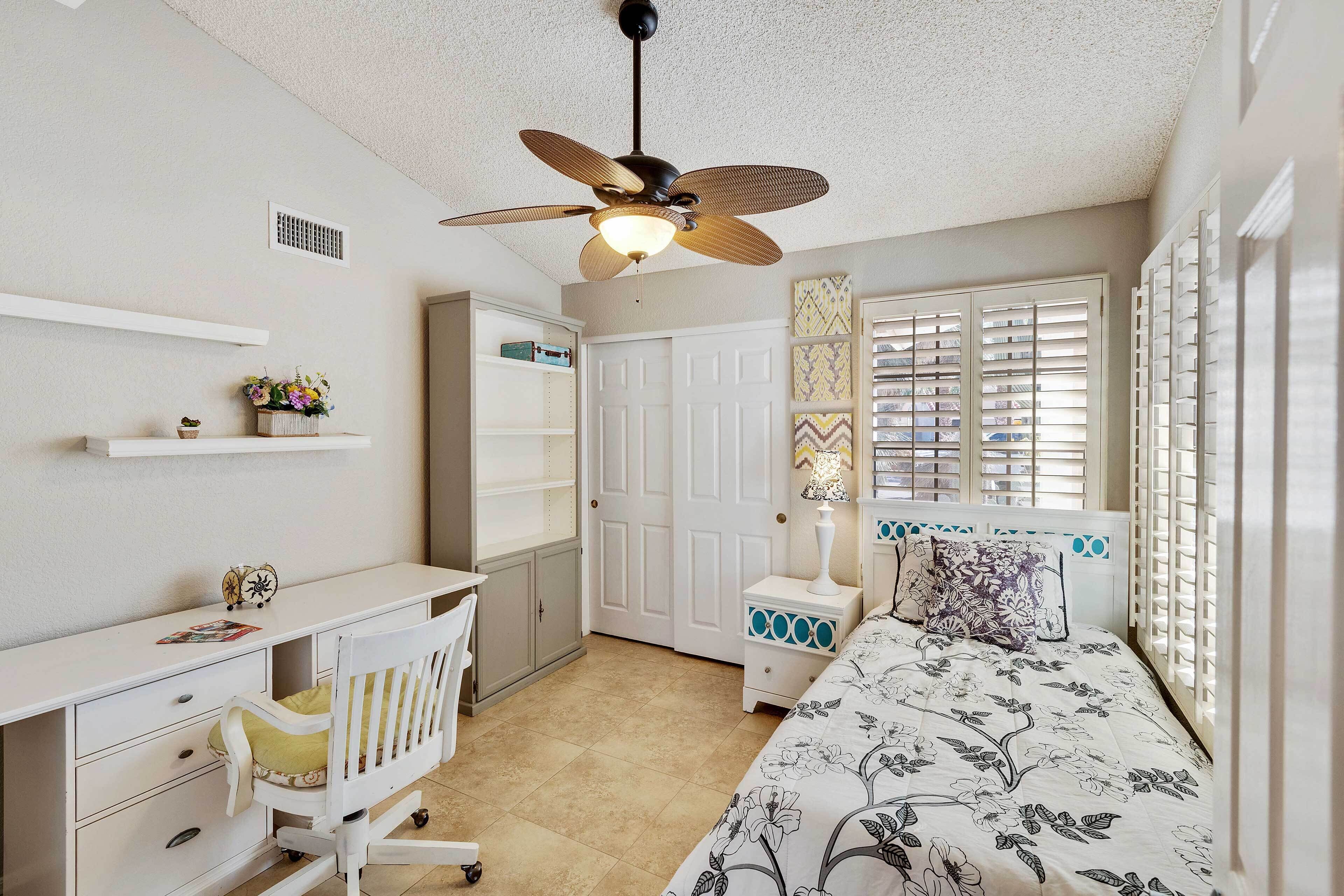 A bedroom with a bed, a desk, a chair, a nightstand, a ceiling fan, and a window with blinds.