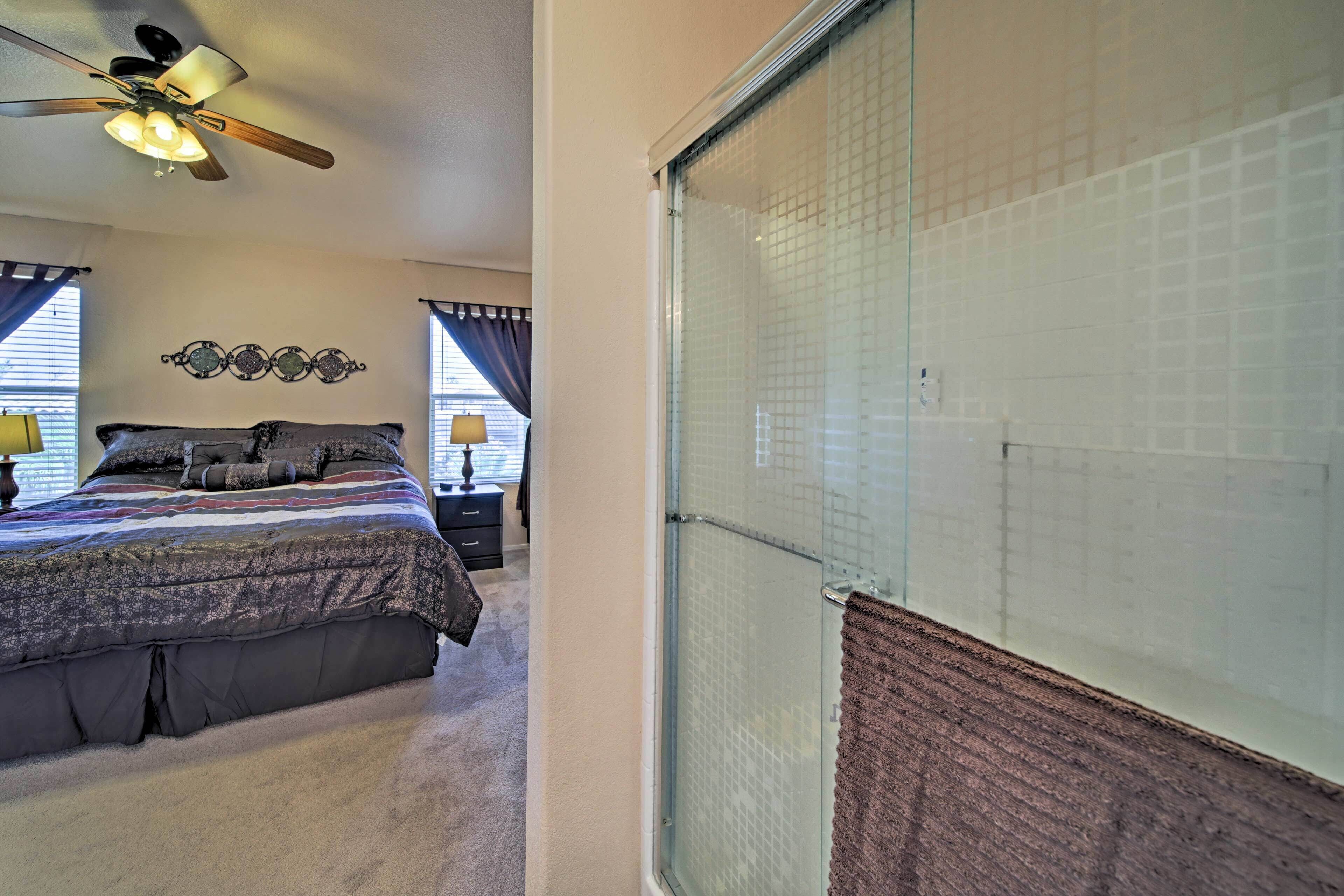 A bedroom with a bed, a ceiling fan, a window with curtains, and a bathroom with a glass shower door.