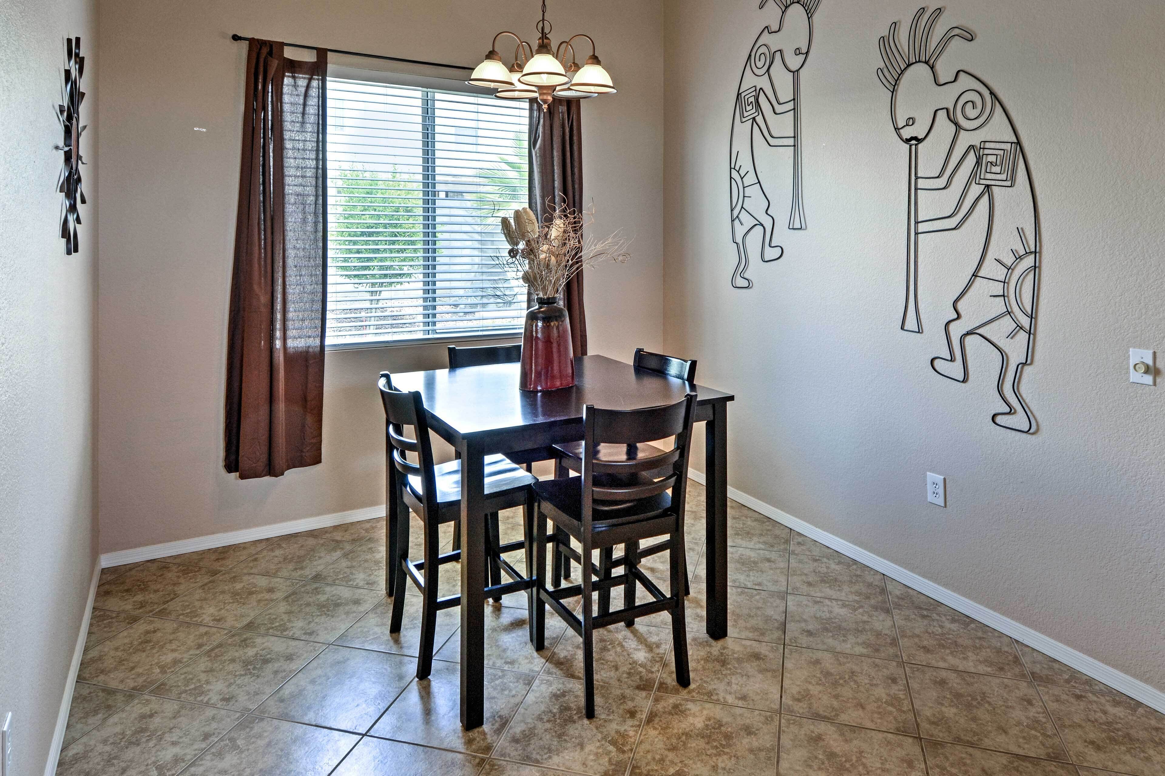 Un comedor con una mesa y sillas, un jarrón con plantas secas, una ventana con cortinas y decoración en la pared.