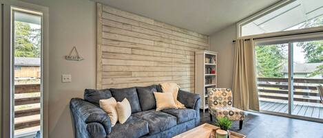 A modern living room with a dark grey sofa, a wooden accent wall, a bookshelf, and a large sliding glass door leading to an outdoor deck.